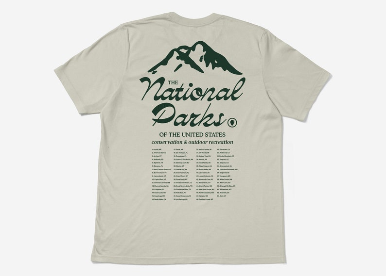 May include: A light beige t-shirt with a green mountain graphic and the words "The National Parks" in a cursive font. Below the text is a list of U.S. National Parks. The shirt promotes conservation and outdoor recreation.