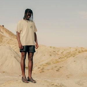 May include: A person wearing a beige t-shirt with a small graphic and black shorts stands in a desert landscape. The t-shirt has a small pocket with a mountain graphic. The person is wearing sandals.