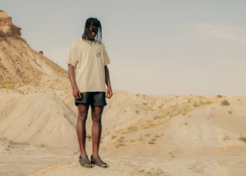 May include: A person wearing a beige t-shirt with a small graphic and black shorts stands in a desert landscape. The t-shirt has a small pocket with a mountain graphic. The person is wearing sandals.