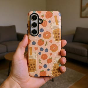 May include: A phone case with a repeating pattern of strawberries, blueberries, donuts, and bubble tea cups on a light peach background. The case has a camera cutout and is held in a hand.