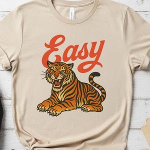 May include: A tan t-shirt with a tiger illustration in orange and black stripes. The word "Easy" is printed above the tiger in orange script. The shirt has short sleeves and a crew neck. The shirt is a neutral colour.