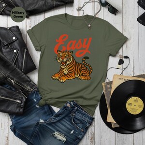 May include: Military green t-shirt featuring the word "Easy" in red script above a tiger illustration. The tiger is orange and black. The shirt is styled with blue denim shorts and a black leather jacket.