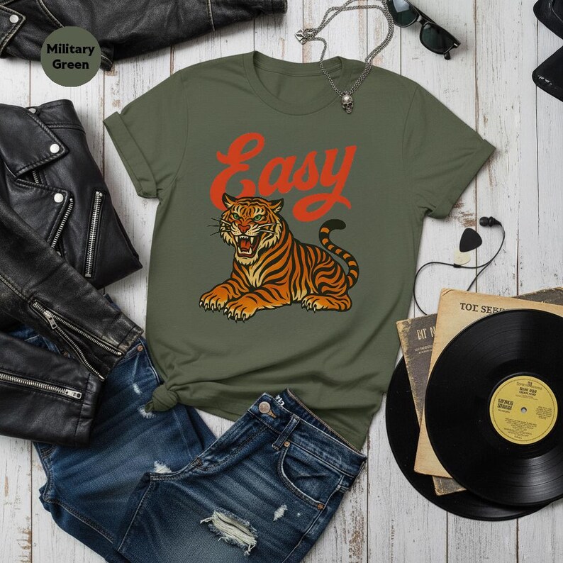 May include: Military green t-shirt featuring the word "Easy" in red script above a tiger illustration. The tiger is orange and black. The shirt is styled with blue denim shorts and a black leather jacket.