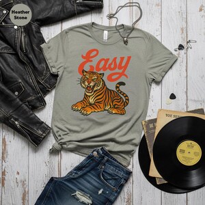 May include: A grey t-shirt with an orange tiger illustration and the word "Easy". The tiger is orange and black. The shirt is styled with blue jeans, a black leather jacket, and vinyl records.
