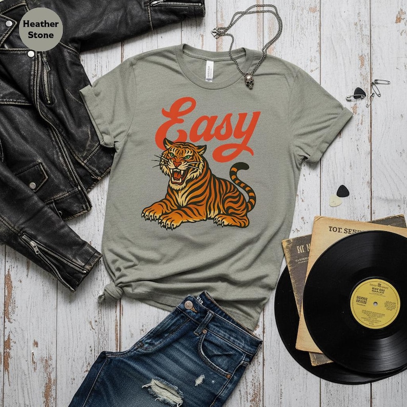 May include: A grey t-shirt with an orange tiger illustration and the word "Easy". The tiger is orange and black. The shirt is styled with blue jeans, a black leather jacket, and vinyl records.