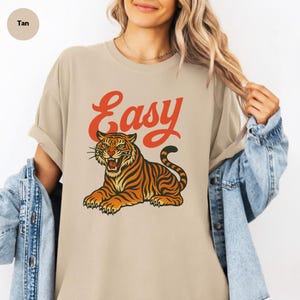 May include: Tan t-shirt featuring the word "Easy" in red script above a tiger illustration. The tiger is orange and black with green eyes, in a relaxed pose. The shirt has a casual, oversized fit.