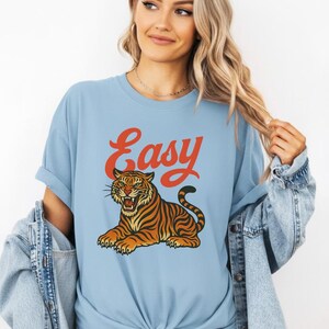 May include: Pale blue t-shirt featuring a tiger illustration and the word "Easy" in orange. The tiger is depicted in orange and black. The shirt has a relaxed fit and rolled-up sleeves.