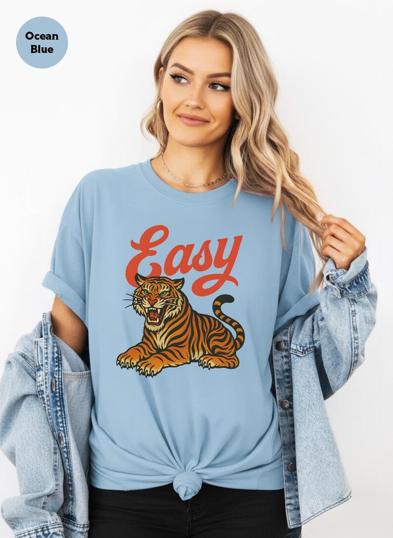 May include: Pale blue t-shirt featuring a tiger illustration and the word "Easy" in orange. The tiger is depicted in orange and black. The shirt has a relaxed fit and rolled-up sleeves.