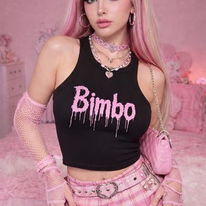 May include: Black crop top with the word "Bimbo" in pink dripping font. The model is wearing a pink plaid skirt, pink accessories, and a pink purse. The background is pink.