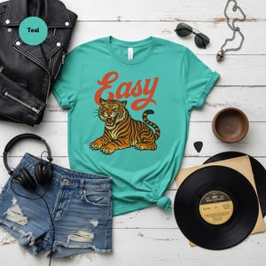 May include: Teal t-shirt with the word "Easy" in orange script and a tiger illustration. The tiger is orange and black with green eyes. The shirt is styled with denim shorts, a black leather jacket, and vinyl records.