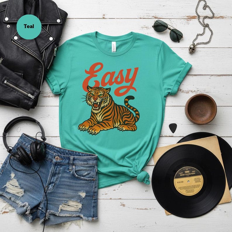 May include: Teal t-shirt with the word "Easy" in orange script and a tiger illustration. The tiger is orange and black with green eyes. The shirt is styled with denim shorts, a black leather jacket, and vinyl records.