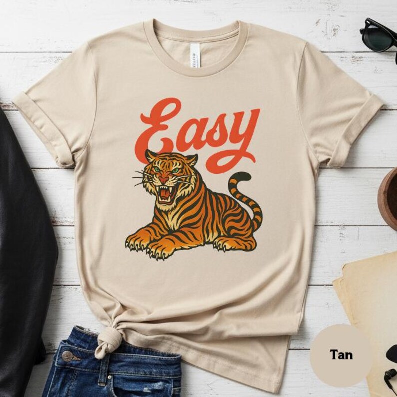 May include: A tan t-shirt with a tiger illustration in orange and black stripes. The word "Easy" is printed above the tiger in orange script. The shirt has short sleeves and a crew neck. The shirt is a neutral colour.