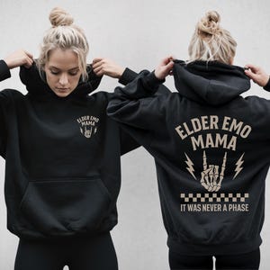 May include: Black hoodie featuring the text "ELDER EMO MAMA" and a skeleton hand making a rock and roll gesture. The phrase "IT WAS NEVER A PHASE" is printed below the graphic. The hoodie has a front pocket and a drawstring hood.
