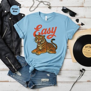 May include: Pale blue t-shirt featuring the word "Easy" in orange script above a tiger illustration. The tiger is orange and black with green eyes. The shirt is styled with denim shorts and a black leather jacket.