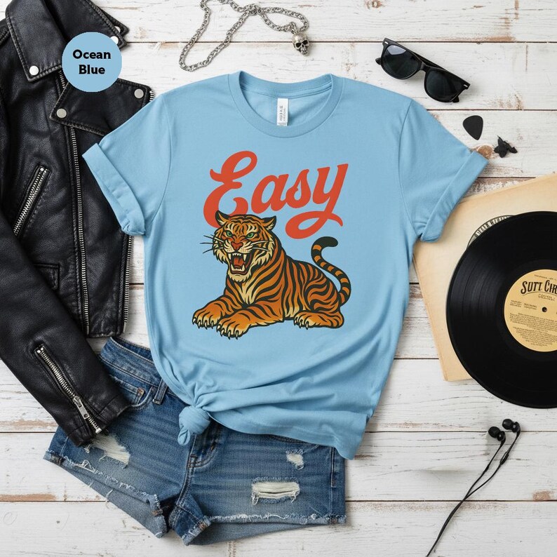 May include: Pale blue t-shirt featuring the word "Easy" in orange script above a tiger illustration. The tiger is orange and black with green eyes. The shirt is styled with denim shorts and a black leather jacket.