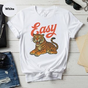 May include: White t-shirt with a retro design of a tiger in orange and black stripes, and the word "Easy" in orange script. The tiger is depicted in a relaxed pose, with its mouth open. The shirt is made of a soft material.