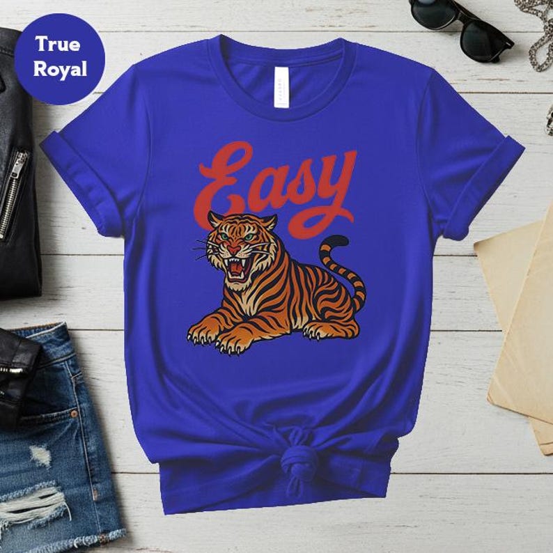 May include: Royal blue t-shirt with a tiger illustration in orange and black stripes, and the word "Easy" in red script. The tiger is lying down with its mouth open. The shirt is knotted at the waist.
