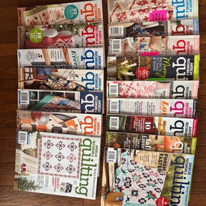 May include: A collection of "American Patchwork & Quilting" magazines, featuring colorful quilt designs and projects. The magazines are stacked on a wooden surface, showcasing various cover designs with text and images related to quilting.