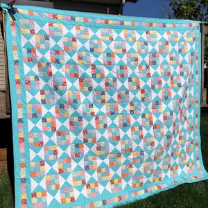 May include: A large, handmade quilt with a turquoise border and backing. The quilt features a repeating pattern of colorful squares and white diamond shapes. The squares contain a variety of floral and geometric patterns in shades of red, orange, yellow, and blue.