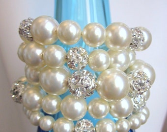 May include: A multi-layered bracelet made of white faux pearls and silver rhinestone embellishments. The bracelet is wrapped around a blue glass vase.