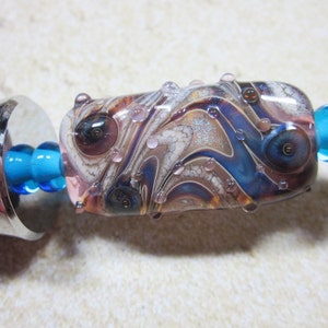 Peut inclure: Gros plan sur une perle de verre color&eacute;e avec des tourbillons de bleu, de brun et de blanc. La perle a une surface textur&eacute;e avec de petites bulles et des paillettes. Elle est attach&eacute;e &agrave; une cha&icirc;ne en m&eacute;tal argent&eacute; avec des perles de verre bleues.