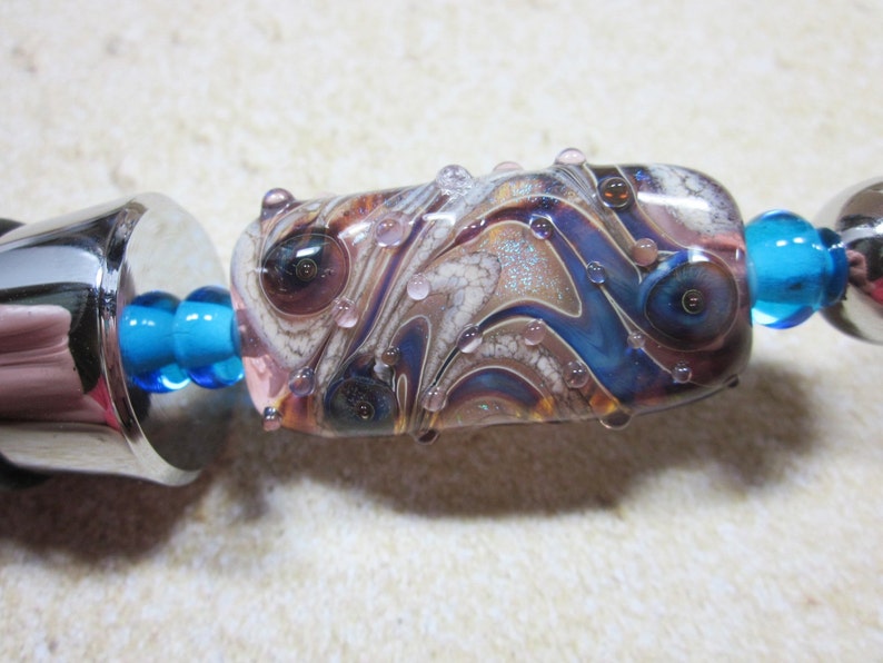 Peut inclure: Gros plan sur une perle de verre color&eacute;e avec des tourbillons de bleu, de brun et de blanc. La perle a une surface textur&eacute;e avec de petites bulles et des paillettes. Elle est attach&eacute;e &agrave; une cha&icirc;ne en m&eacute;tal argent&eacute; avec des perles de verre bleues.