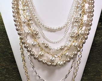 May include: A multi-strand necklace featuring a mix of gold and silver chains, white and cream pearls, and clear crystal beads.