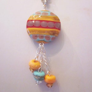 May include: A silver chain necklace with a round, multi-colored glass pendant. The pendant has a pattern of gold, red, and blue stripes with small pink dots. The pendant is attached to three smaller beads, one blue and two orange.
