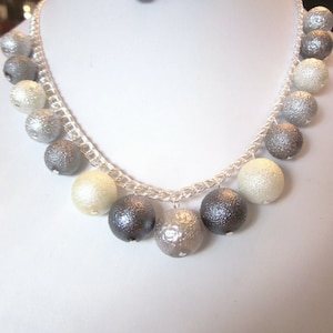 Puede incluir: Un collar de cadena de plata con una hebra graduada de cuentas grises y blancas. Las cuentas son redondas y tienen una superficie texturizada. El collar se combina con pendientes a juego.