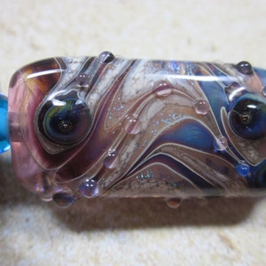 Peut inclure: Une perle en verre avec un motif tourbillonnant de violet, blanc et bleu. La perle a de petites bulles de verre transparentes &agrave; la surface.