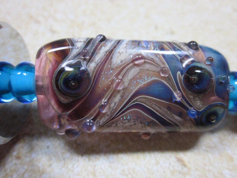 Peut inclure: Une perle en verre avec un motif tourbillonnant de violet, blanc et bleu. La perle a de petites bulles de verre transparentes &agrave; la surface.