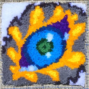 May include: A square, handmade rug with a vibrant eye design. The central eye is a bright blue with a green center, surrounded by purple and yellow accents. The background is gray, with a white grid visible at the edges. This unique rug adds a pop of color.
