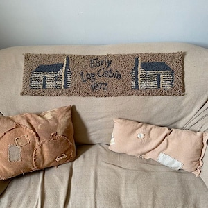 May include: A brown rug with two cabin illustrations and the text "Early Log Cabin 1872". Two decorative pillows in shades of tan and peach are on a beige sofa.