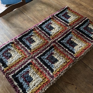May include: A rectangular, hand-crocheted rug with a geometric pattern. The rug features squares with a log cabin design in shades of red, orange, yellow, blue, black, and cream. The rug is placed on a wooden table.