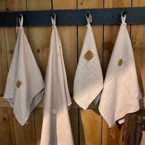 May include: Four off-white linen towels hanging on a dark blue wooden rack. Each towel has a small, irregularly shaped hole near the center. The towels are suspended against a backdrop of vertical wooden planks.
