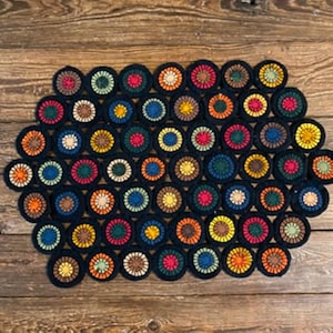 May include: A handmade, colorful crochet rug with a dark blue background. The rug is composed of numerous circular motifs in various colors, including red, orange, yellow, and green. The rug is placed on a wooden surface.