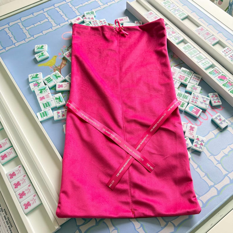 Puede incluir: Una bolsa de terciopelo rosa intenso con cord&oacute;n y una cinta rosa a juego atada en forma de cruz en la parte delantera. La cinta lleva impreso el texto THE MERRY MAHJONG CO en un patr&oacute;n repetitivo. La bolsa tiene una textura suave y afelpada.