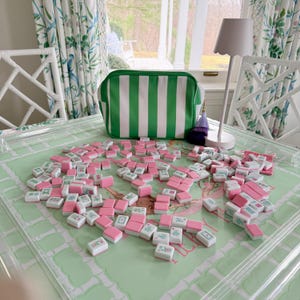 May include: A collection of pink and white Mahjong tiles scattered on a green patterned surface. A green and white striped bag sits behind the tiles. A white lamp and chair are also in the background.
