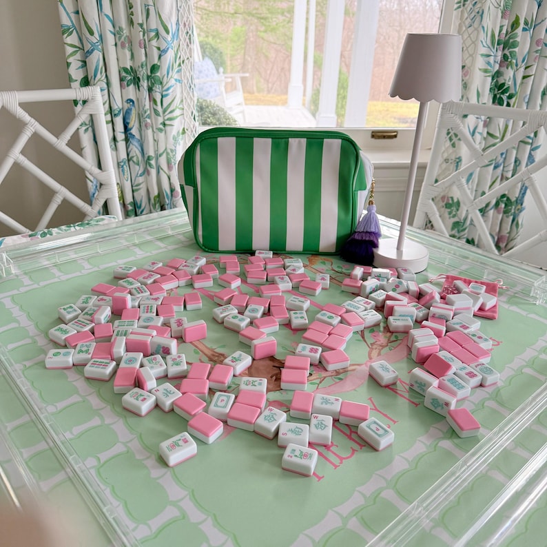 May include: A collection of pink and white Mahjong tiles scattered on a green patterned surface. A green and white striped bag sits behind the tiles. A white lamp and chair are also in the background.
