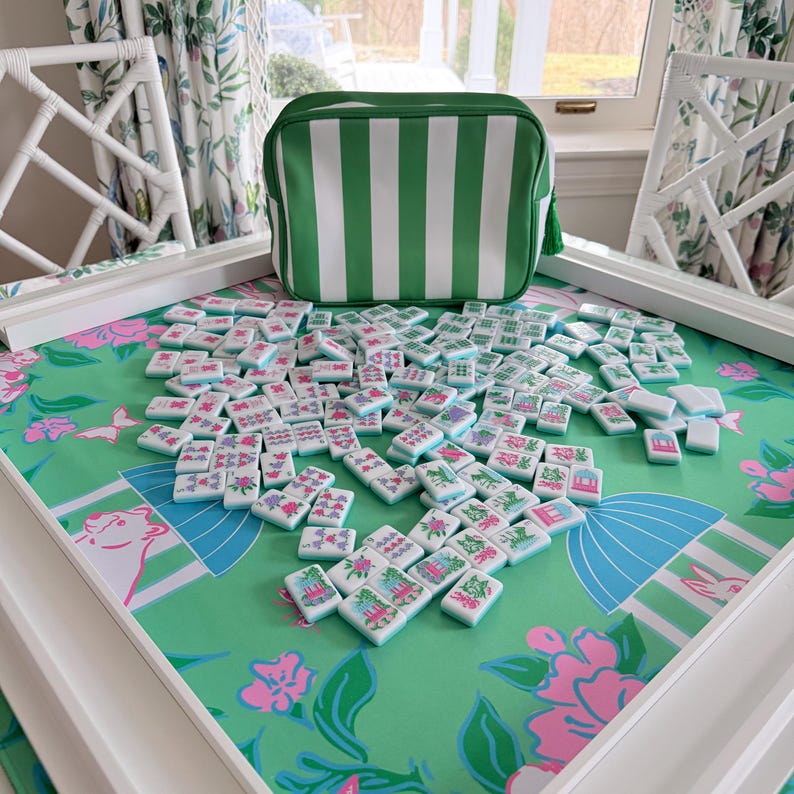 May include: A mahjong set with white tiles featuring colorful illustrations, scattered on a green floral patterned surface. A green and white striped carrying case sits nearby. The set is displayed on a white table with matching chairs.