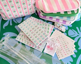 May include: A mahjong set featuring white tiles with floral and character designs, clear acrylic racks, a small white drawstring bag with two pink dice, and instructional cards. Includes a floral print pouch and several striped zippered cases.