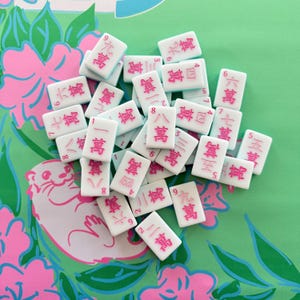 May include: A collection of white Mahjong tiles with pink characters and numbers. The tiles are scattered on a green and pink floral patterned surface. The game pieces are rectangular and feature various Chinese characters and numbers.