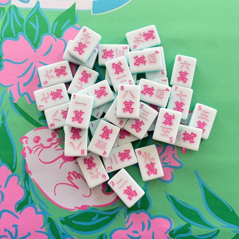 May include: A collection of white Mahjong tiles with pink characters and numbers. The tiles are scattered on a green and pink floral patterned surface. The game pieces are rectangular and feature various Chinese characters and numbers.