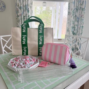 May include: A cream-colored tote bag with green handles and the word "Mahj" printed on the straps. A pink and white striped cosmetic bag, a white tray filled with tiles, and a clear acrylic tile rack are also visible.