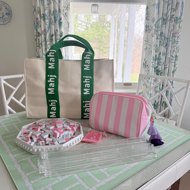 May include: A cream-colored tote bag with green handles and the word "Mahj" printed on the straps. A pink and white striped cosmetic bag, a white tray filled with tiles, and a clear acrylic tile rack are also visible.