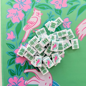 May include: A collection of white mahjong tiles with green and blue markings and numbers. Some tiles feature a pink bird illustration. The tiles are scattered on a green background with pink floral and bird patterns.