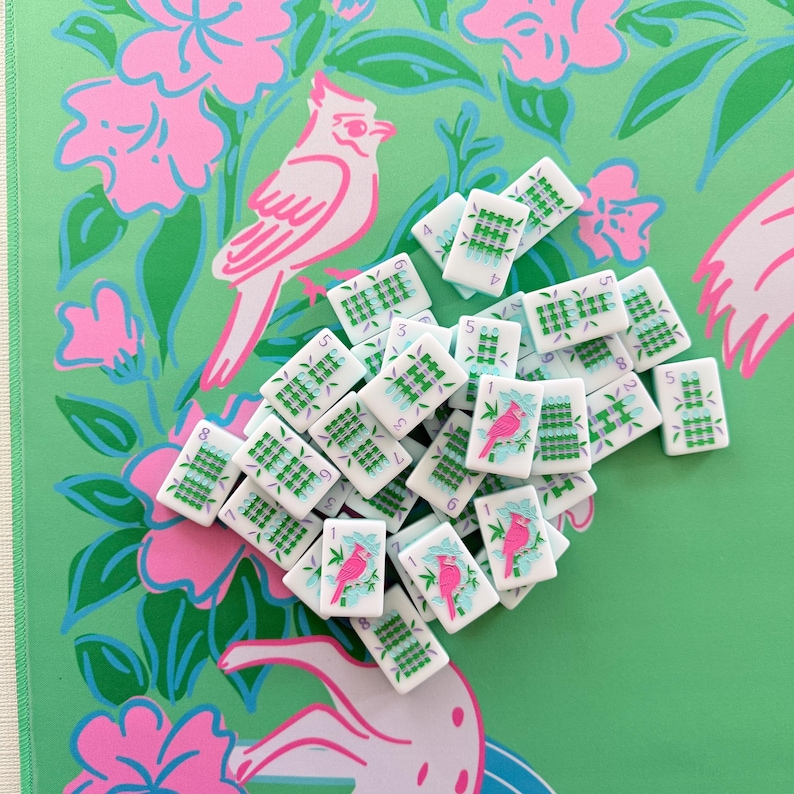 May include: A collection of white mahjong tiles with green and blue markings and numbers. Some tiles feature a pink bird illustration. The tiles are scattered on a green background with pink floral and bird patterns.