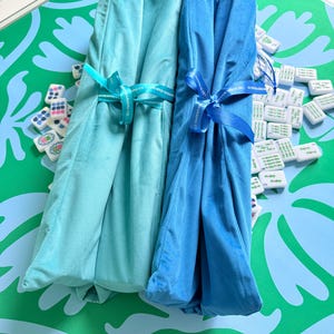 Puede incluir: Dos tapetes de tela plegados en tonos turquesa claro y azul brillante, cada uno asegurado con una cinta a juego impresa con el texto The Merry Mahjong Co. Los tapetes est&aacute;n colocados sobre una superficie con patr&oacute;n verde y azul claro con fichas.