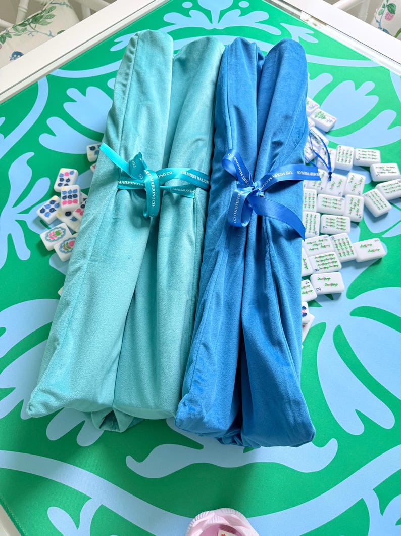 Puede incluir: Dos tapetes de tela plegados en tonos turquesa claro y azul brillante, cada uno asegurado con una cinta a juego impresa con el texto The Merry Mahjong Co. Los tapetes est&aacute;n colocados sobre una superficie con patr&oacute;n verde y azul claro con fichas.