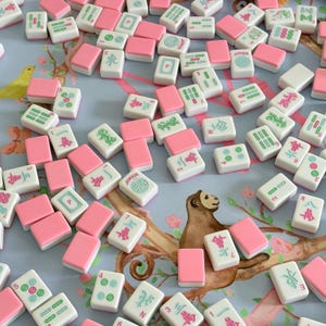 May include: A collection of Mahjong tiles scattered on a floral patterned surface. The tiles are white with pink accents and feature various symbols and numbers. Some tiles have the word "Joker" printed on them. The game pieces are ready to play.